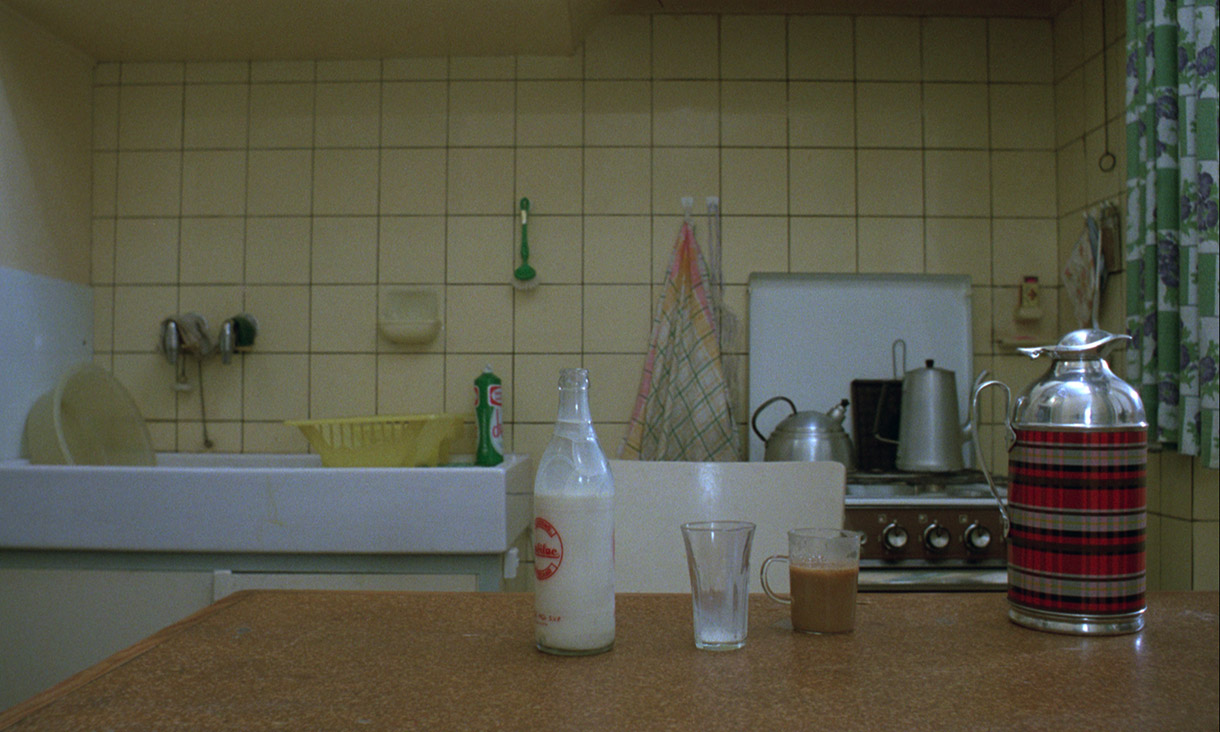 A bottle of milk and two glasses are on a bench in a kitchen