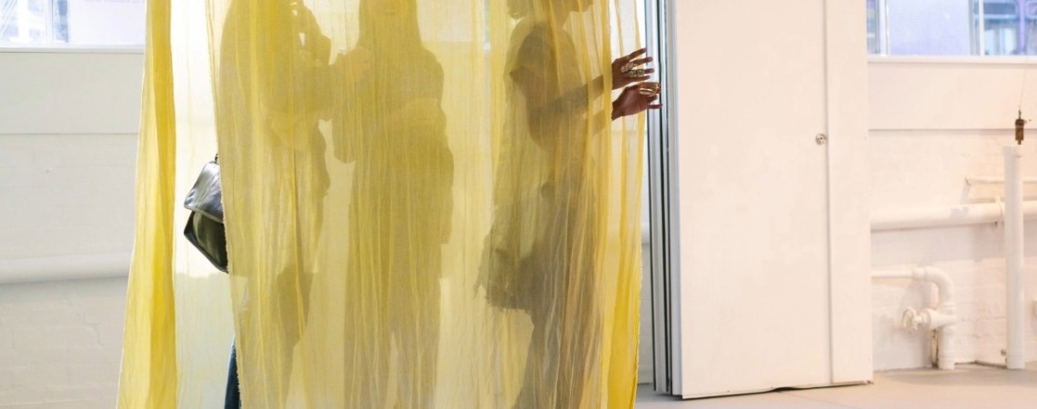 Three people shrouded i nyellow curtains. Two hands, wearing several rings, emerge as they open the curtains. 