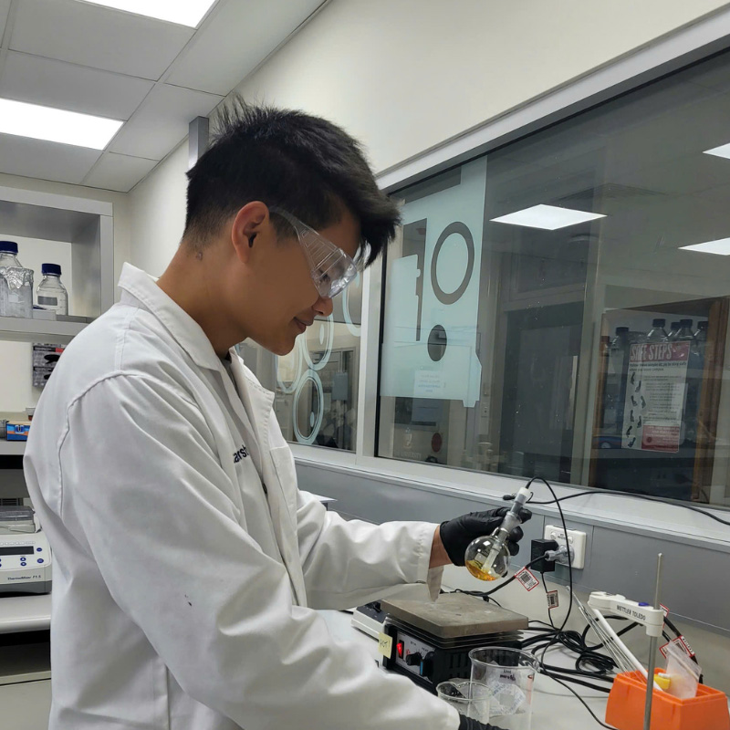 Dr Hao in a lab coat and black glove conducting an experiment in a laboratory, operating scientific equipment on a bench with beakers and a flask.