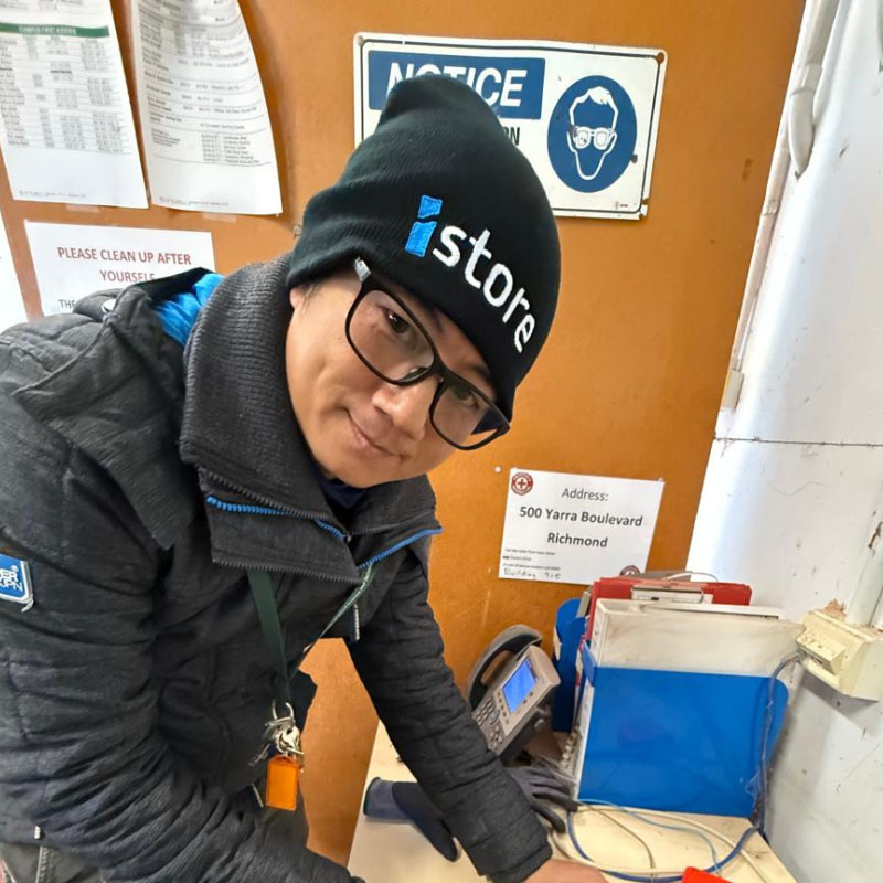 Dr Khantam in a dark winter jacket and beanie leaning over a desk in an office with safety notices and papers on the wall, surrounded by office supplies.
