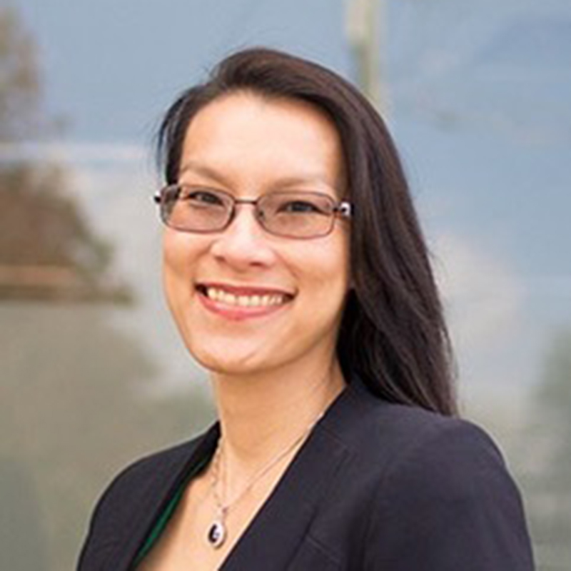 Photograph of Eleanor Mak. She smiles directly at the camera. She is has long, dark brown hair, wears glasses and wears a black blazer.