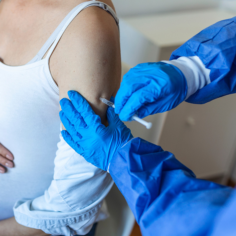 Person receiving a vaccine injection in their upper arm from a healthcare worker wearing blue gloves and protective clothing.