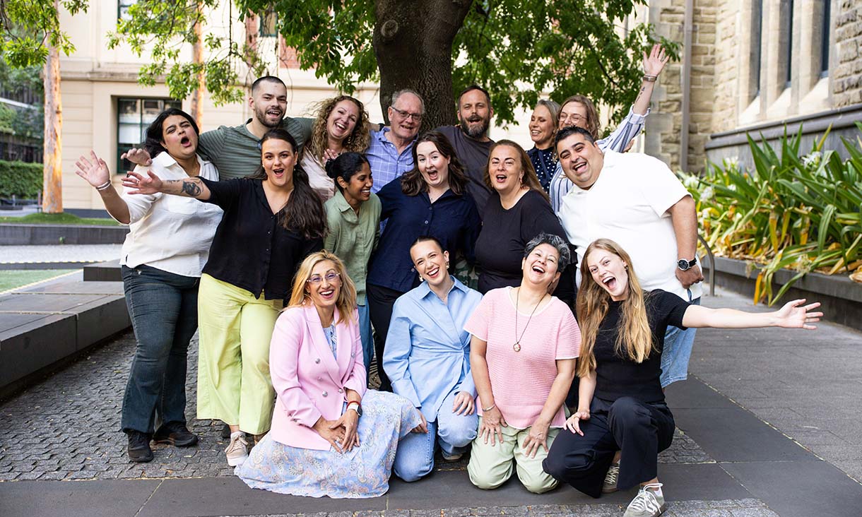 Group of people posing together outdoors under a tree, some standing and others kneeling, with arms raised and smiling, in front of campus buildings.