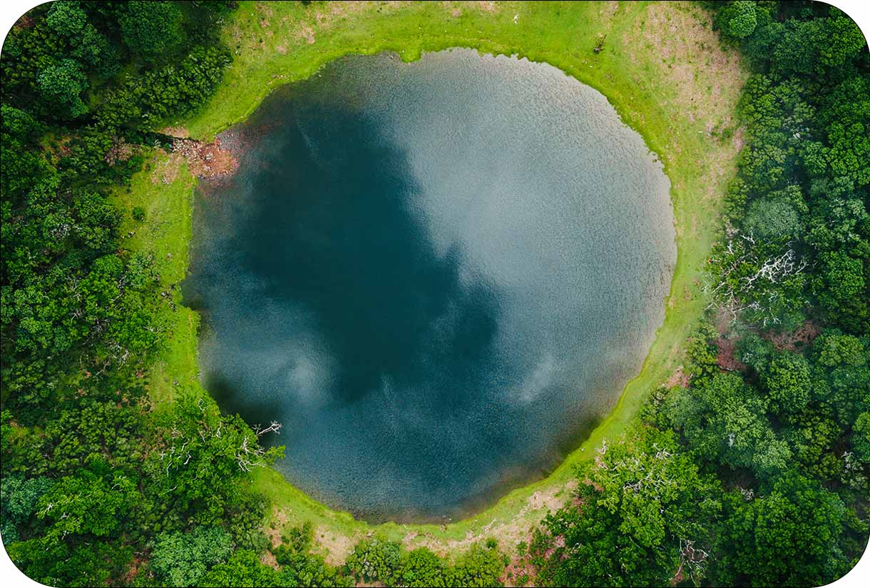 Aerial view of a round lake surrounded by dense green forest and grassy edges.