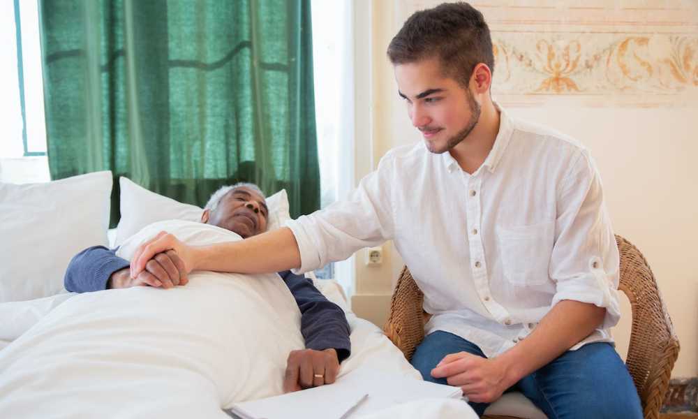 Young person sitting bedside of elderly person in bed and holding their hand