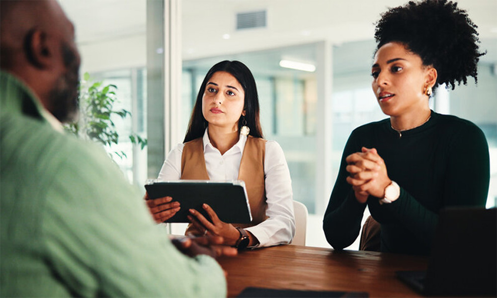 Three people sitting at a table in a modern office, engaged in a professional discussion with a digital tablet.