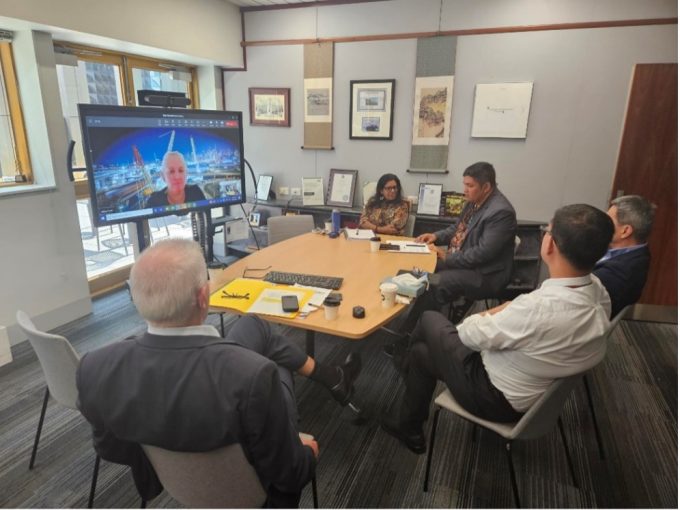 Five people sitting around a table in a meeting with a screen showing another participant.