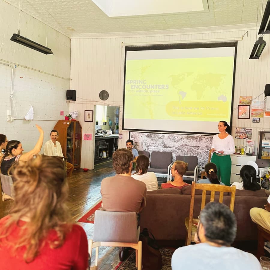 A group of seated adults attend a presentation in a large room in an old building. The presenter stands near a large screen displaying a slide titled “Spring Encounters: 2023 with(in) place – This is how we do Praxis in Latin America.”