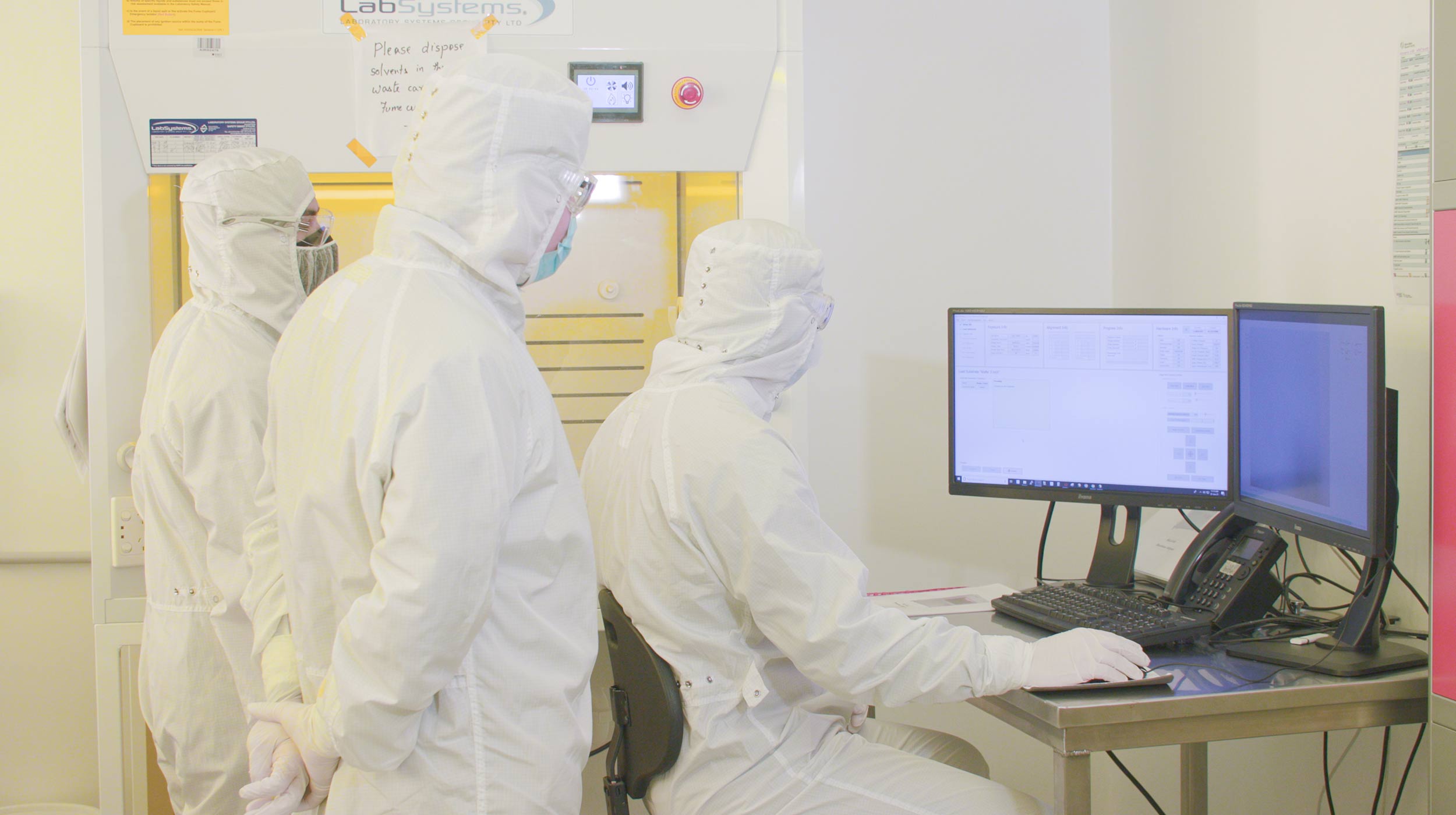 Three people in white protective clothing work on a computer