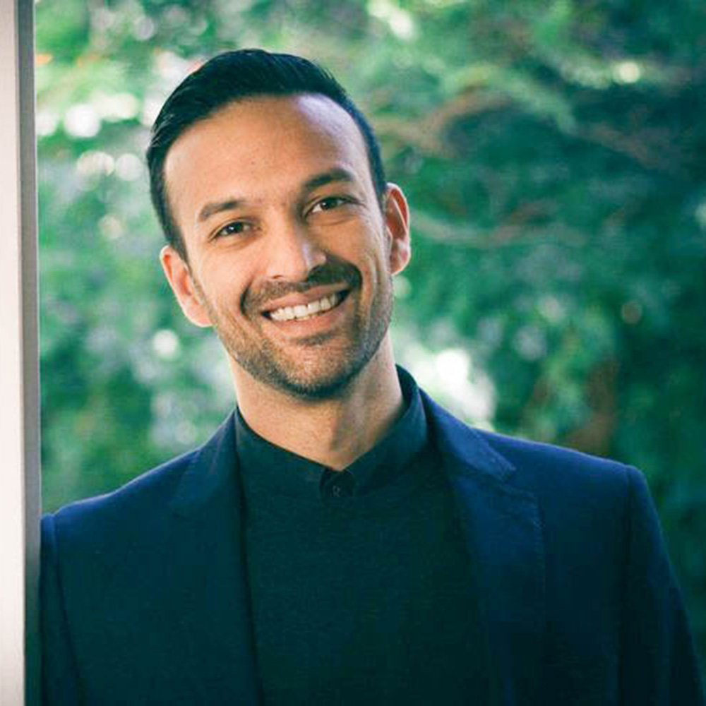 Photograph of Janak Rogers. He wears a black blazer and shirt and is smiling at the camera. His hair is neatly combed and kept out of his face.
