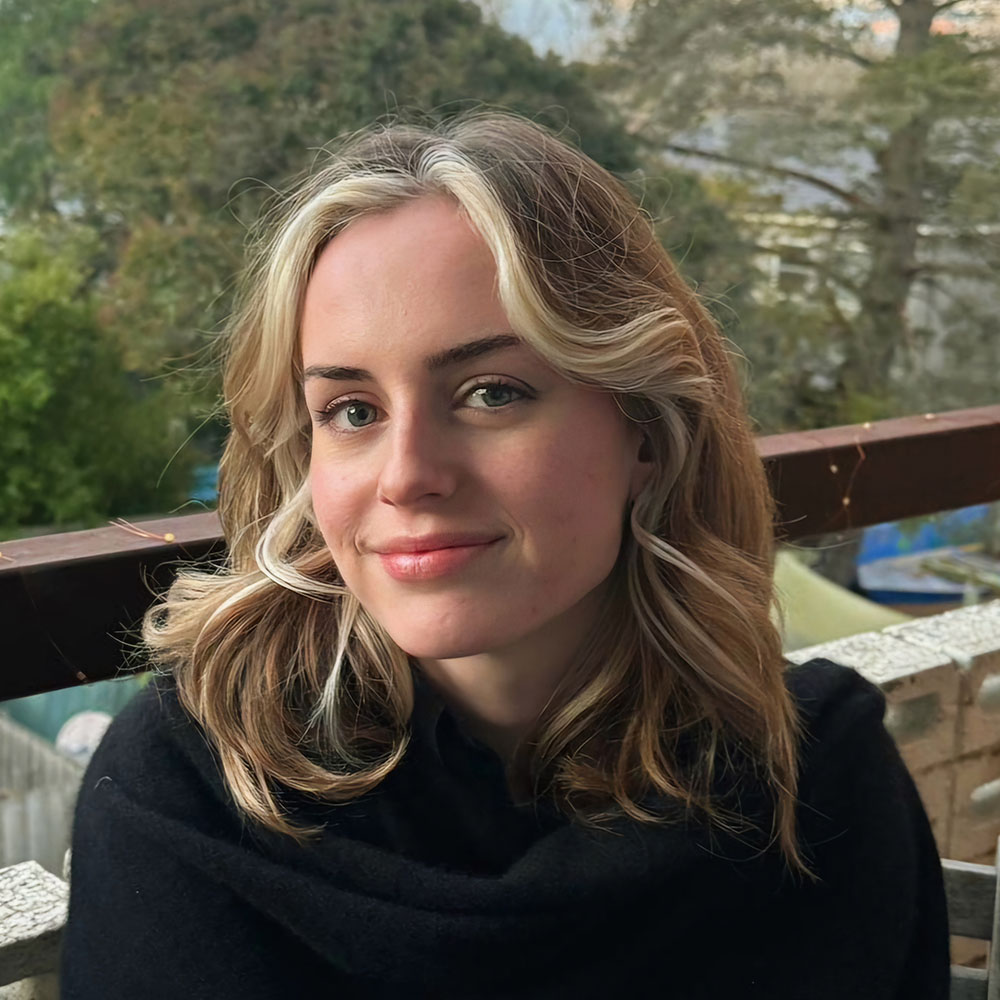 Photograph of Josie Banner. She wears a black jumper and is smiling at the camera with her head slightly tilted away. Her blond hair is out and falls over her shoulders in soft waves.