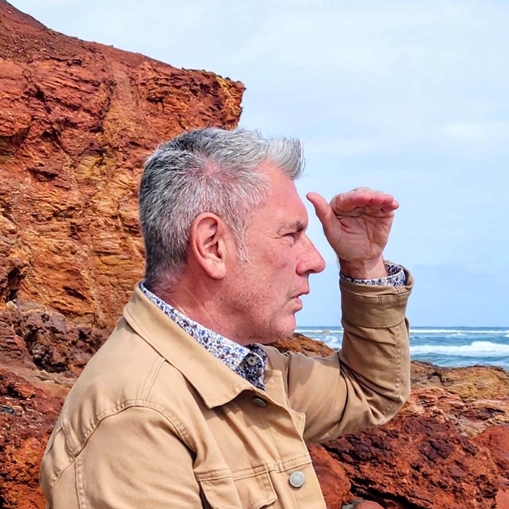 Photograph of Sean Redmond. He faces away from the camera with his hand raised above his eyes. He has short, grey hair and wears a tan-coloured jacket. In the background there is a brown, rocky formation and ocean waves crashing in the distance.