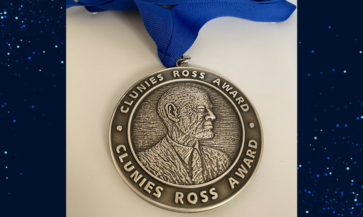 Photo of a metal, circular medal. The medal is attached to a blue ribbon. The medal has the wording "Clunies Ross Award' written twice around the circumference - separated by two dots. In the centre is the profile of a man in a formal shirt and tie.