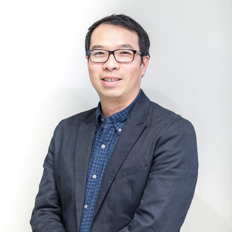 Profile photo of Professor Alan Wong. Alan is standing at an angle and smiling at camera in front of a grey wall.