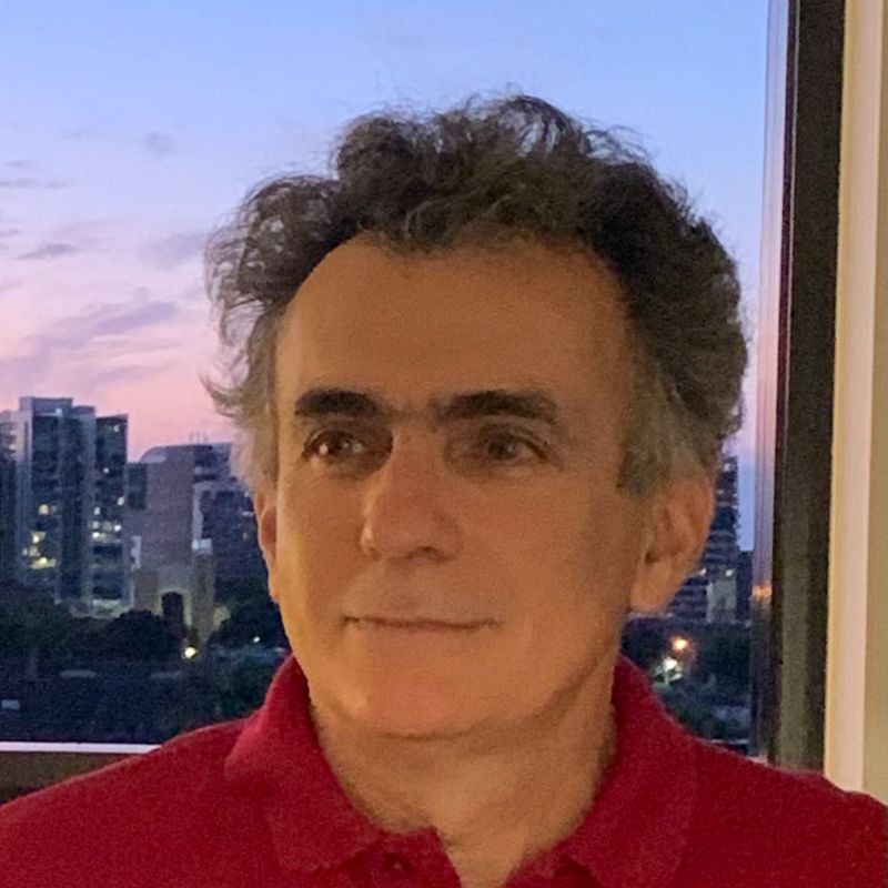 Profile photo of Professor Alireza Bab-Hadiashar. Alireza is standing in front of a window, standing straight on to the camera and looking off to the side. Outside the window is a cityscape with a sunset sky (pink to blue gradient). Multi-story buildings can be seen in the distance with some floors having their lights on.