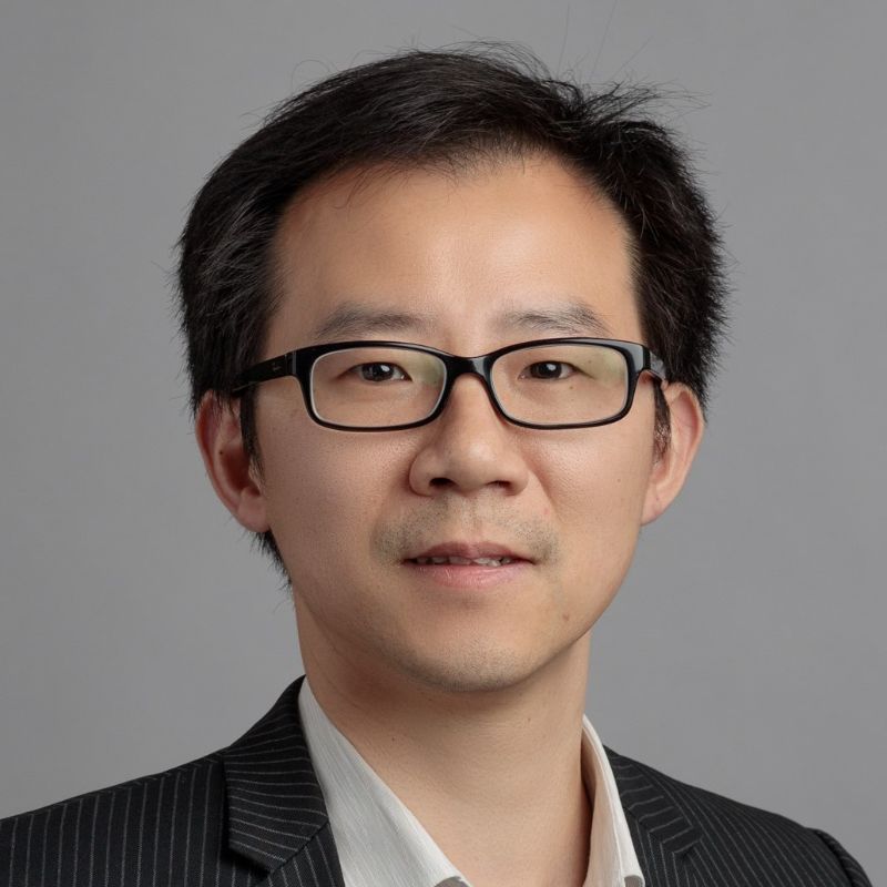 Profile photo of Dr Annan Zhou. Annan is standing in front of a grey wall. Annan is wearing a suit shirt, blazer and glasses. Annan is looking at the camera and smiling.