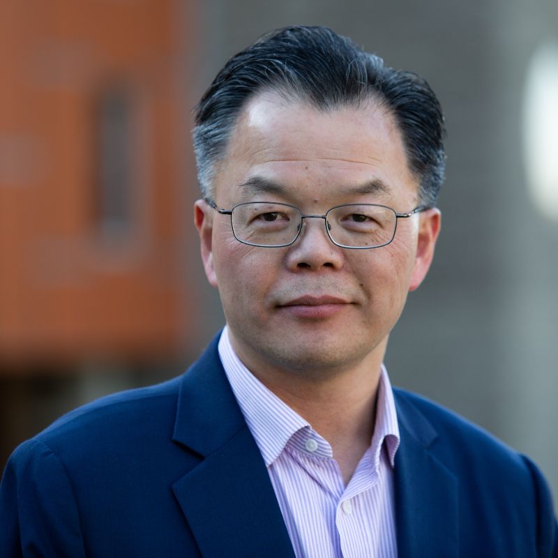 Profile photo of Professor Charlie Xue. Charlie is standing outside, the background is out of focus but appears to be of a building corner - one side painted maroon, the other grey. Charlie is standing at an angle, lookinjg at the camera and smiling. Charlie is wearing glasses, a button up shirt and blazer.