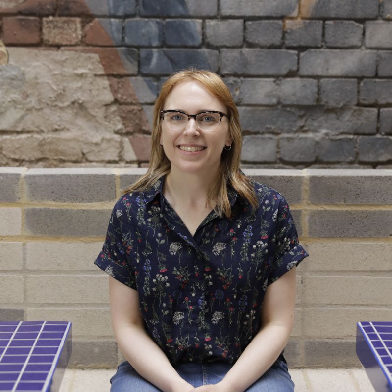 Grace Mcquilten profile photo. She sits in front of a painted brick wall while wearing glasses and smiling.