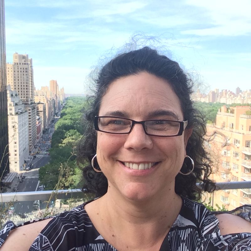 Profile photo of Dr Jenny Robinson. Jenny is standing on an elevated area. Behind Jenny is a cityscape filled with tall buildings and trees. The photo shows Jenny from shoulders up. Jenny is looking at the camera and smiling.