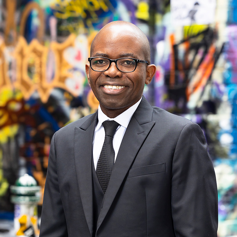 Portrait of Mathews Nkhoma standing in a city laneway in front of graffiti artwork