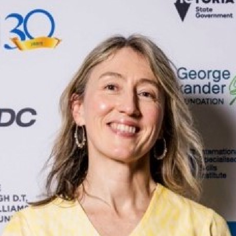 Portrait of Melanie Kyle smiling towards the camera against a media wall. Melanie has shoulder length light coloured hair and is wearing a yellow blouse.