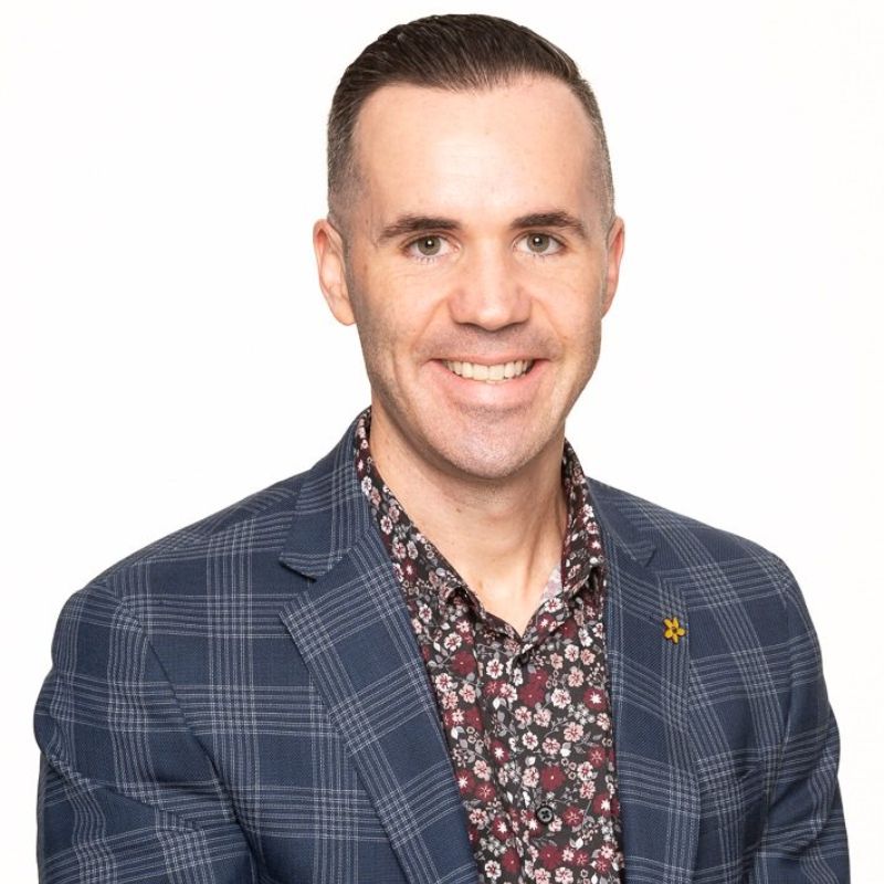 Profile photo of Nick Patterson. Completely white background. Photo is taken from Nick's shoulders to the top of Nick's head. Nick is standing at a slight angle to the camera, looking at the camera and smiling. Nick is wearing a flower-patterend button up shirt and a navy grid patterend blazer with a flower on Nick's right lapel. 