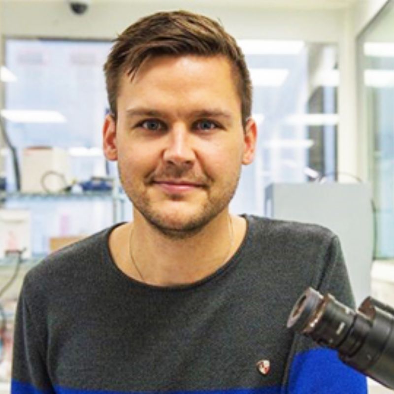 Profile photo of Torben Daenake in a lab, smiling towards the camera. Torben is wearing a blue and grey striped shirt.