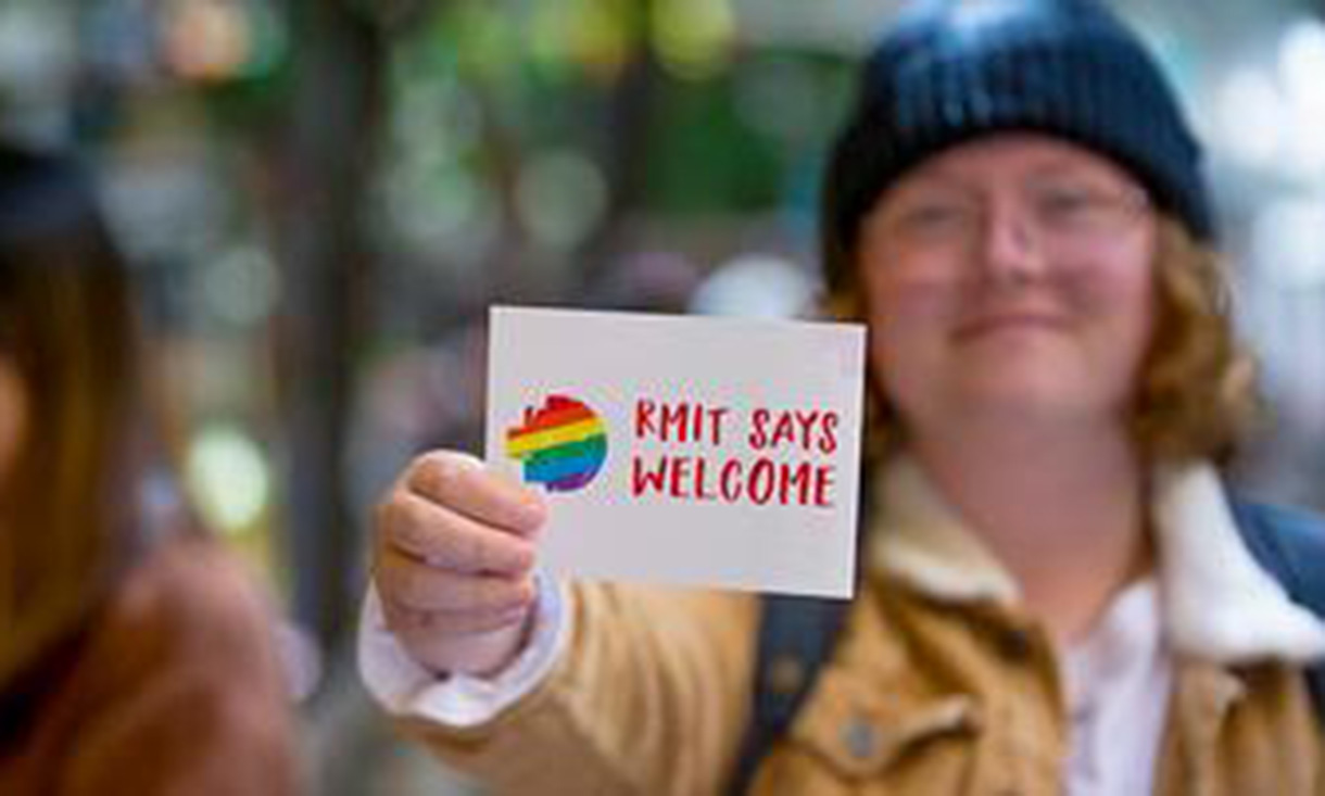A person smiling and holding a sign that reads ‘Pride Safe Welcome,’ symbolising inclusivity for the LGBTQIA+ community