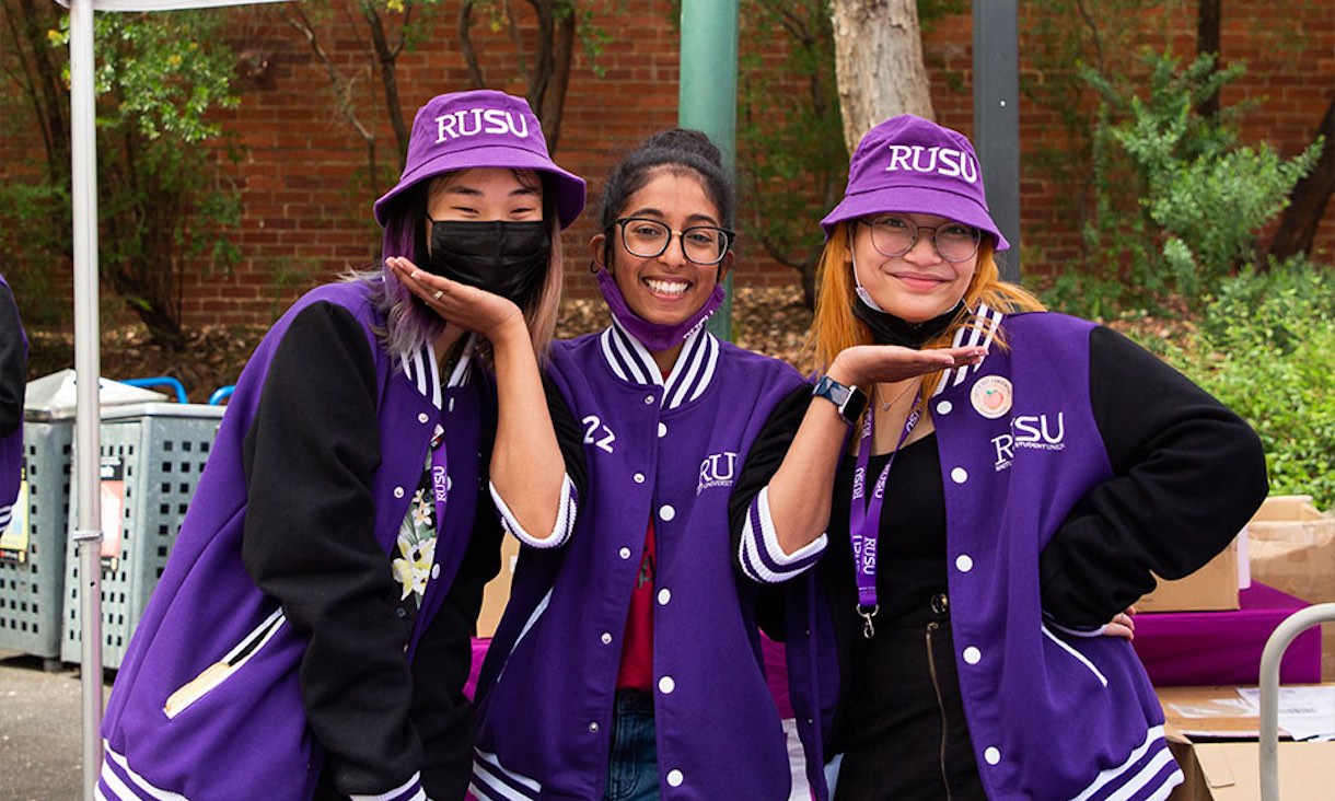 Three RUSU volunteers wearing purple jackets smile at the camera.
