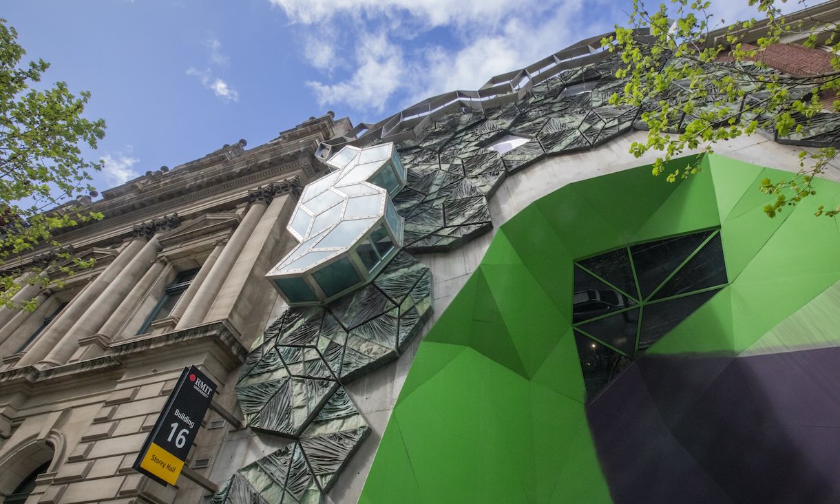 Photo showing the geometric green and glass facade of RMIT Building 16, Storey Hall, blending modern architectural elements with the historic exterior under a partly cloudy sky, with tree branches in the foreground.