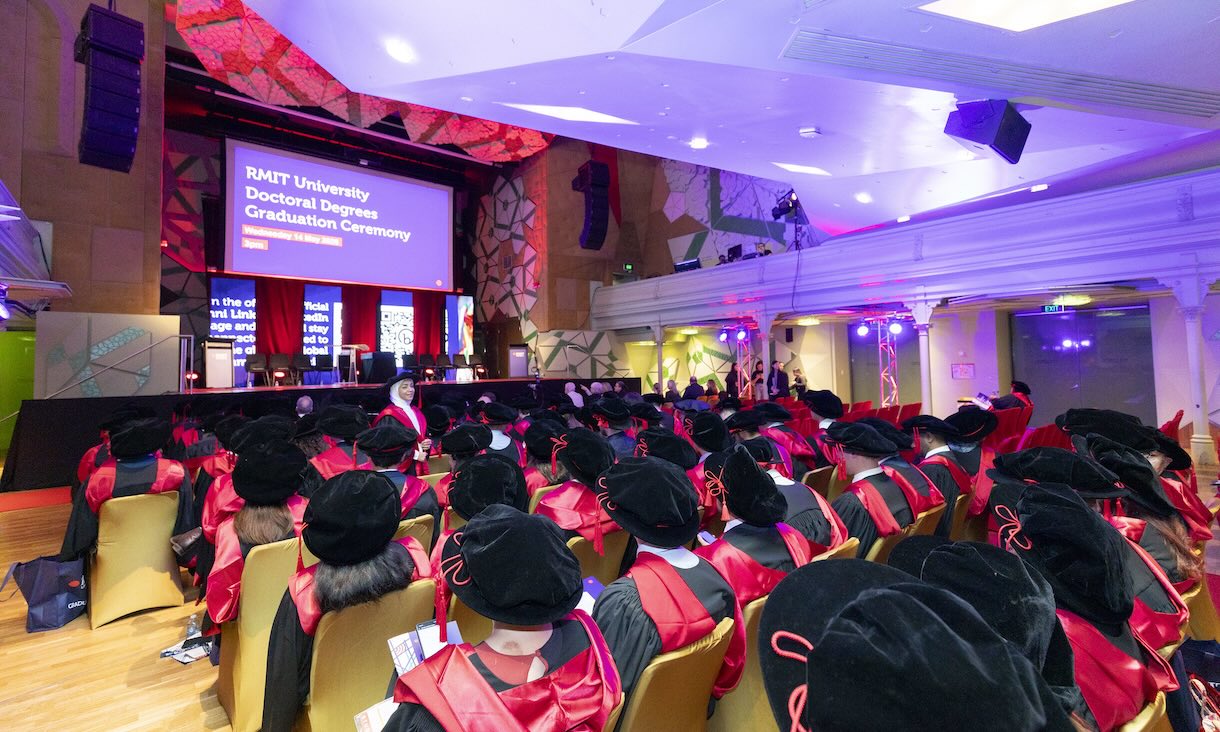 Female graduate looking at camera and holding a flag in front of people and a doughnut van on Bowen Street