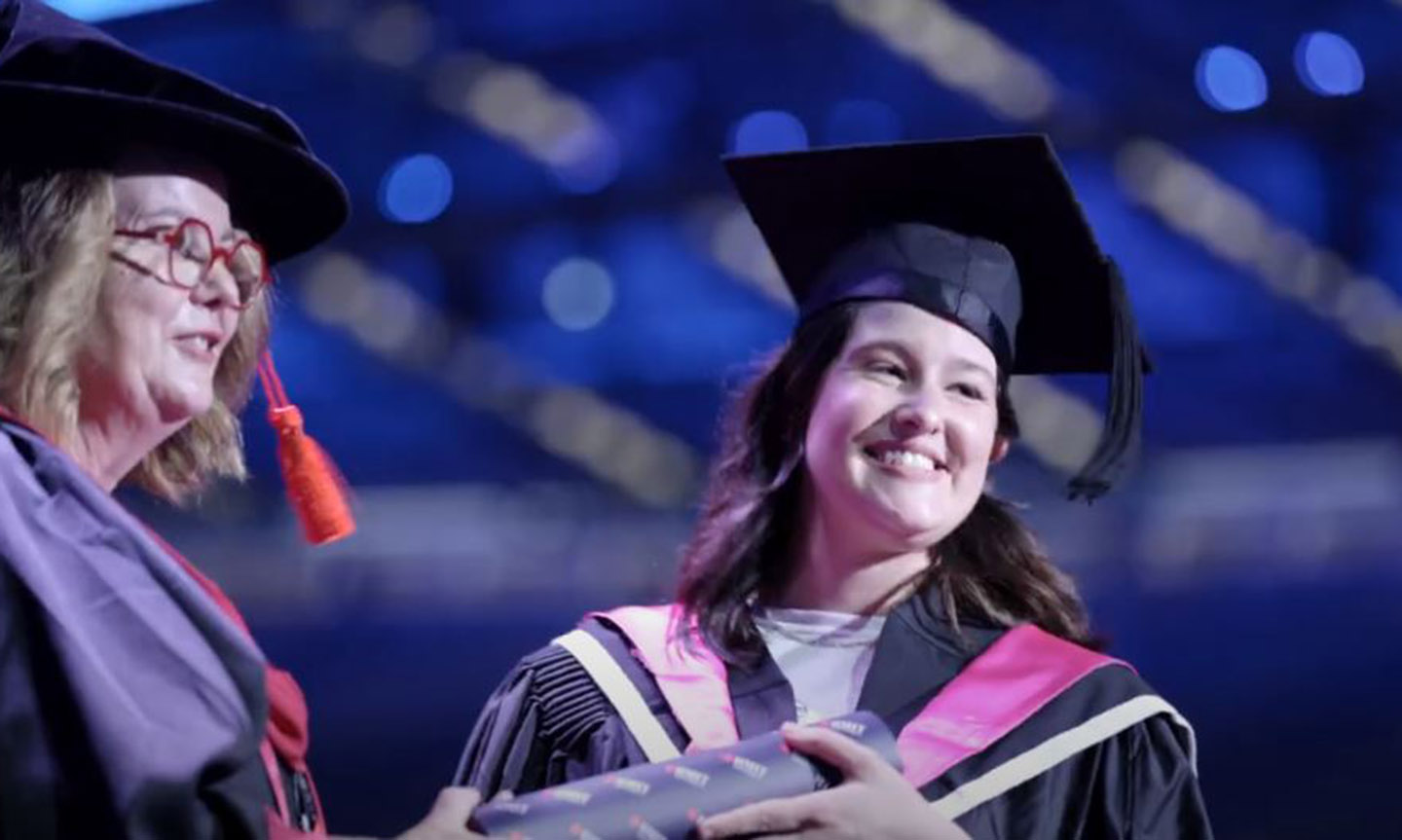A female graduate in academic dress smiles as she receives her graduation scroll from a female academic on stage at the ceremony.