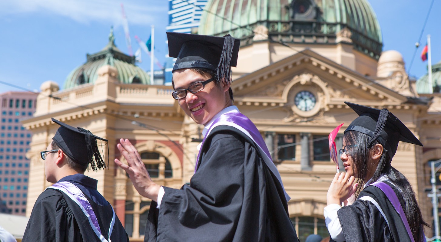 A graduating student wearing black and purple robes waves in front of Flinders Street Station.