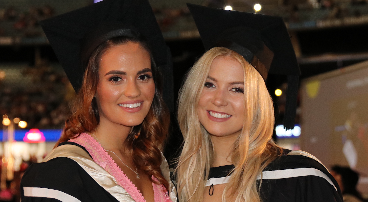 Two students in black and while graduation gowns smile at the camera. They're at the Melbourne Graduation Ceremony.