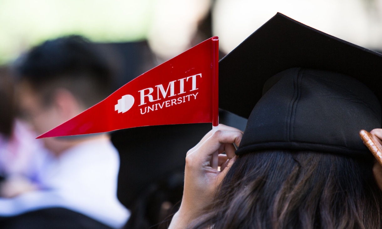 A student holding an RMIT flag adjusts her graduation mortar board hat.