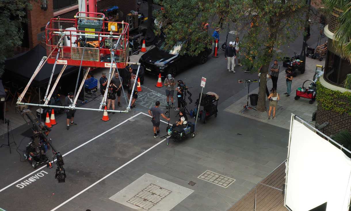 A crane hangs over Bowen Street with film crew walking underneath it with cameras and equipment