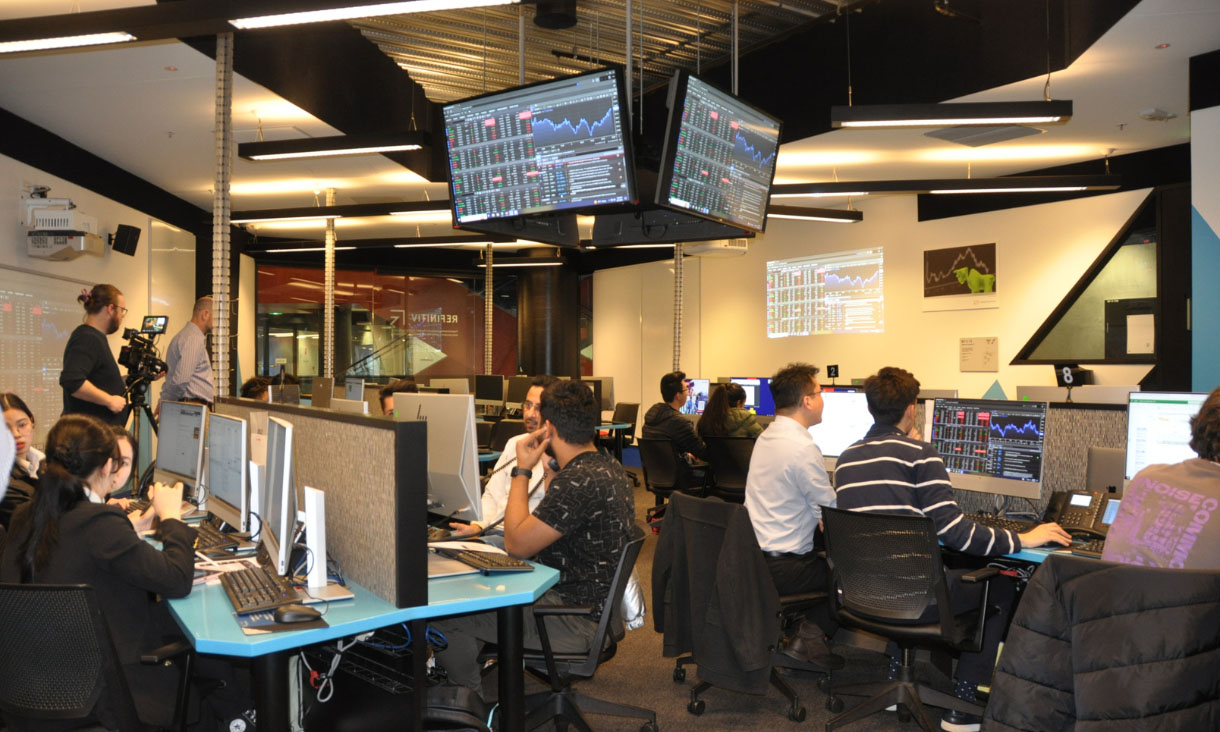 Students sitting at computers with trading information on screens hanging from the ceiling
