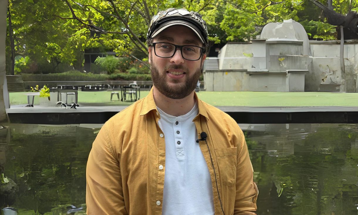 Photography of RMIT student Ben Lambert wearing a black hat and yellow shirt standing smiling towards camera with greass and university campus in background 