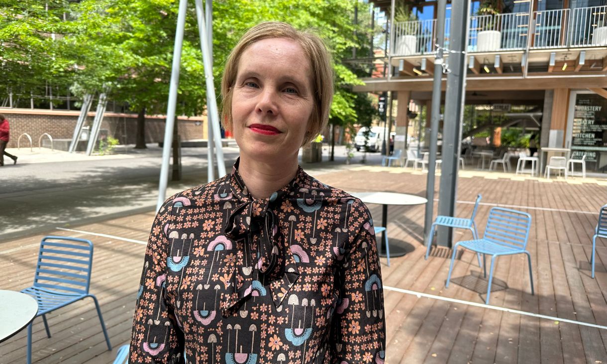 Photography of Associate Professor Brigid Magner standing outside on Bowen Street at RMIT City Campus near tables and chairs with trees in the background. 
