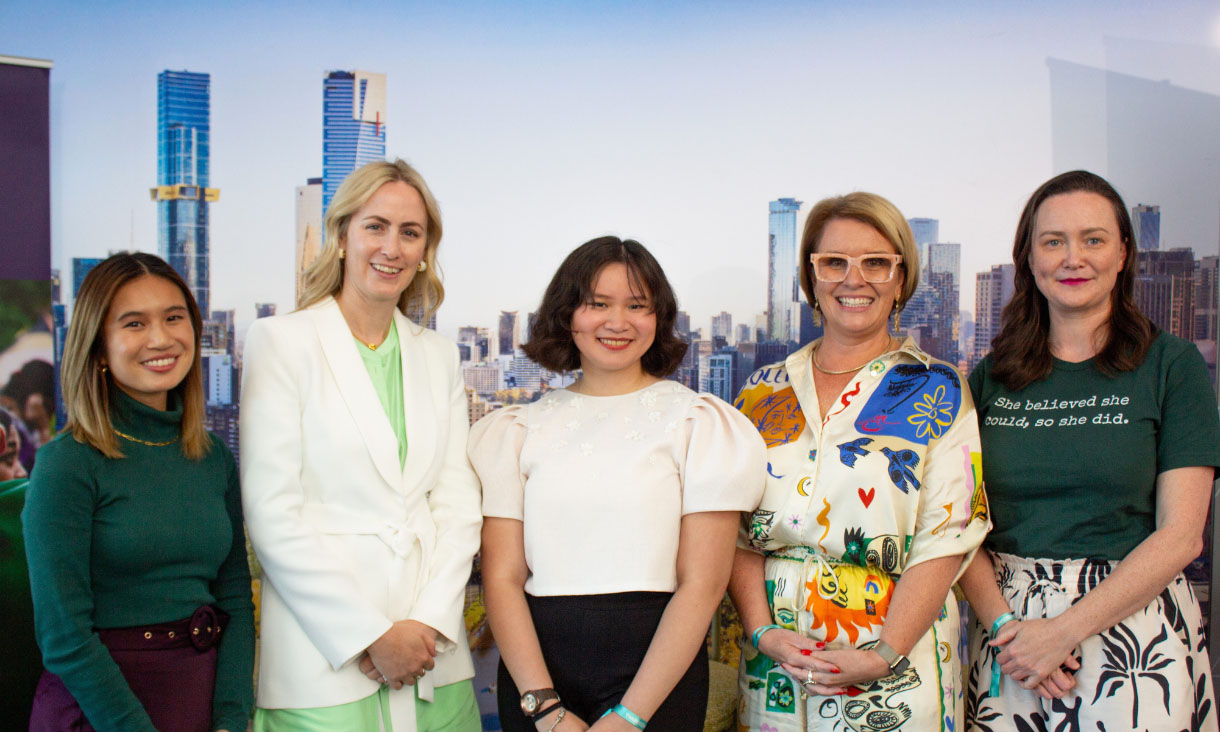 Five women stand together, posing in front of an image of the Melbourne skyline. 