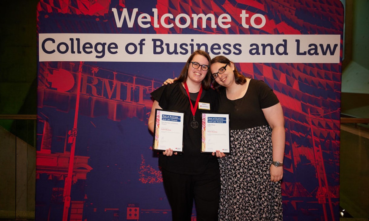 Two women stand in front of a media wall reding ‘Welcome to College of Business and Law’, holding two certificates.