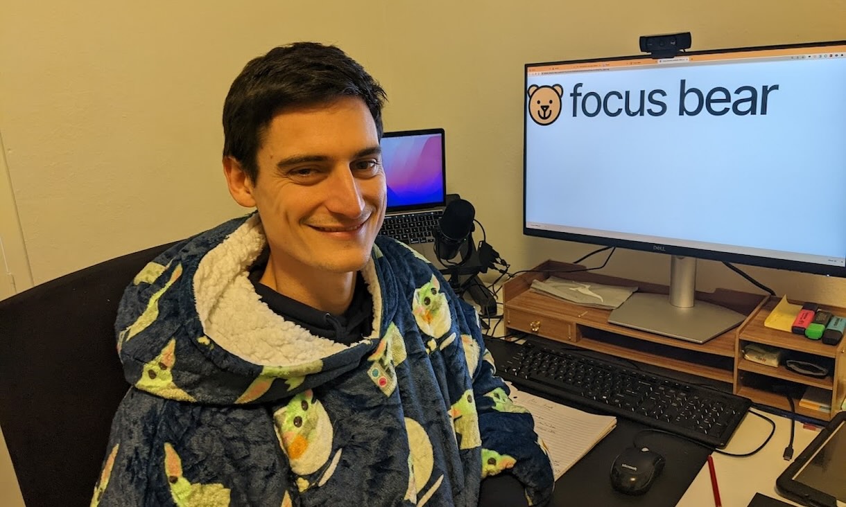 A person wearing a blue hooded blanket with avocado prints sits at a desk with a monitor displaying the Focus Bear logo, a laptop, microphone, keyboard, mouse, notebook, stationery, and other office items.