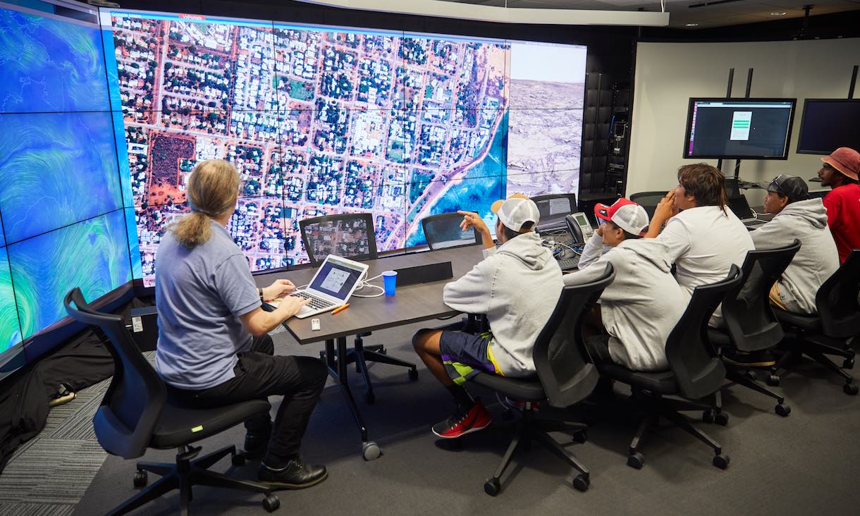 Students sit in front of a large screen displaying arial photography