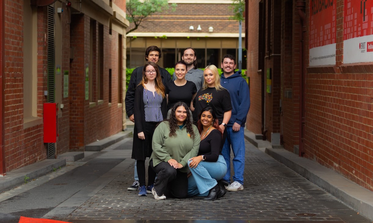 A group of eight people posing together in a laneway lined with brick buildings, featuring casual attire and an urban campus background with RMIT signage visible on the right.