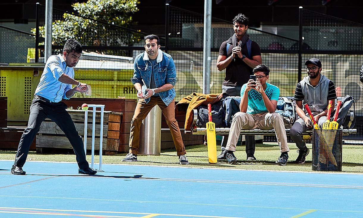 A student prepares to hit an oncoming cricket ball while four students watch in the background