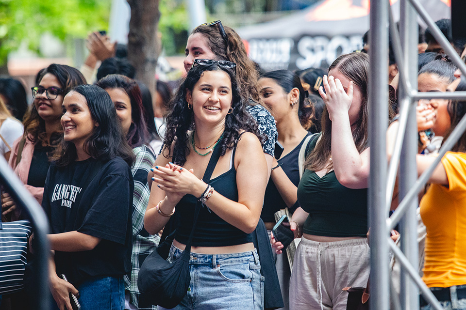 Girl with dark curly hair and wearing a black singlet top stands in a crowd and smiles