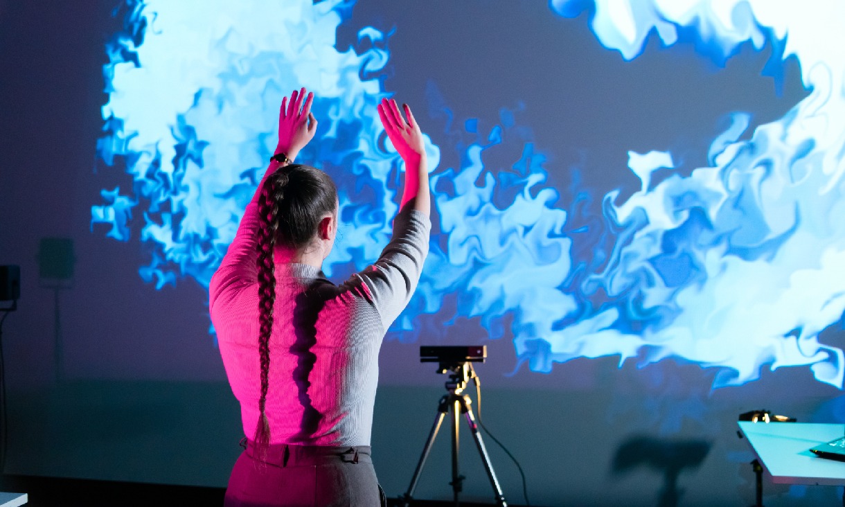 A student standing in front of a motion tracking camera and a screen that shows ripples that follow their movements