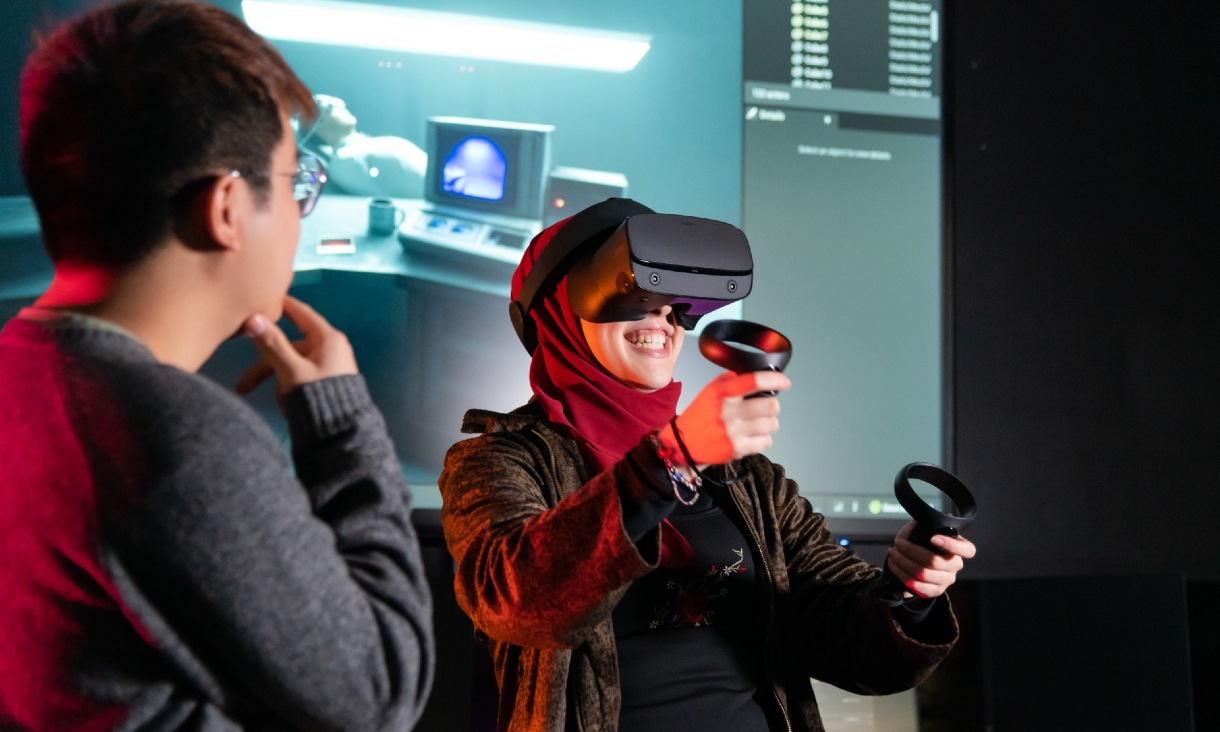 A game design student using a VR headset with handheld controllers while another student watches