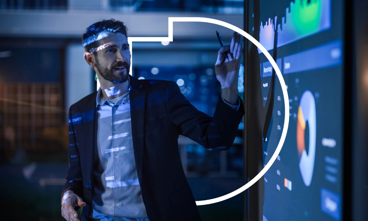A man pointing to charts and graphs on a digital screen during a presentation in a dimly lit room.