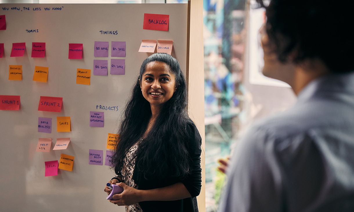Marketing professional standing in front of kanban wall while smiling to colleague 